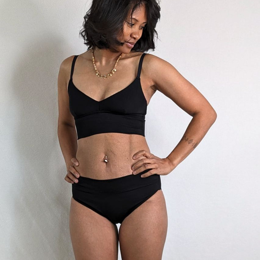 Le modèle appuie ses mains sur ses hanches de et pose dans un ensemble Emma composé de Bralette et d'un slip.