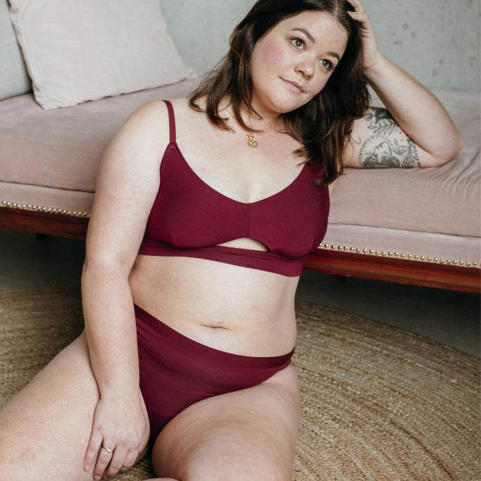 Le modèle est assis sur le sol et s'appuie sur un canapé rose dans le Thea rouge Bralette et Slip.