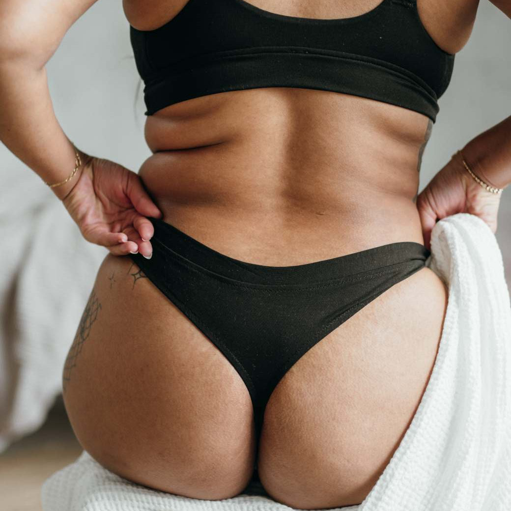 La modella è seduta su una sedia con una coperta. Si vede la schiena, il String nero e il Bralette.