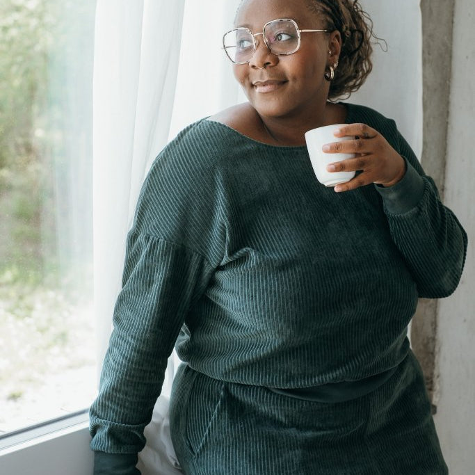 Model trägt jadegrünes Pyjama und steht am Fenster. Sie sitzt halb auf dem Fenstersims, eine Tasse Kaffee in der Hand.