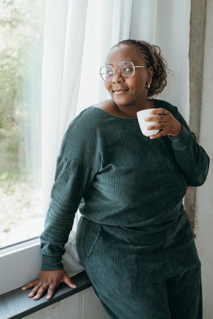 Model trägt jadegrünes Pyjama und steht am Fenster. Sie sitzt halb auf dem Fenstersims, eine Tasse Kaffee in der Hand.