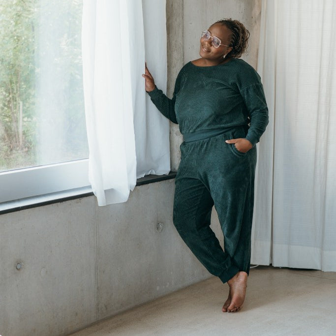 Model trägt jadegrünes Pyjama in Cord und steht am Fenster. Sie öffnet den Vorhang und lächelt hinaus.