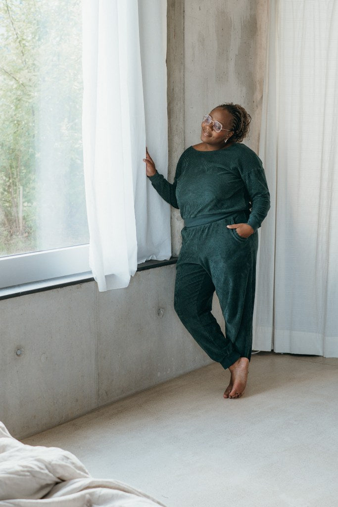 Model trägt jadegrünes Pyjama in Cord und steht am Fenster. Sie öffnet den Vorhang und lächelt hinaus.