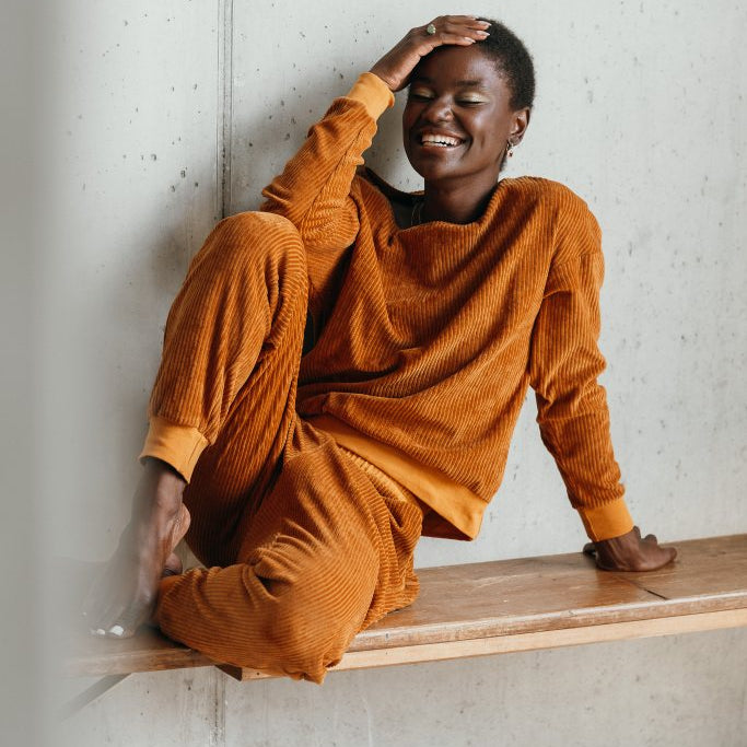 Model trägt ockerfarbigen Pyjama. Sie sitzt auf einer Sitzbank, die Beine auf der Bank angewinkelt und stützt den Kopf auf dem Ellbogen ab.