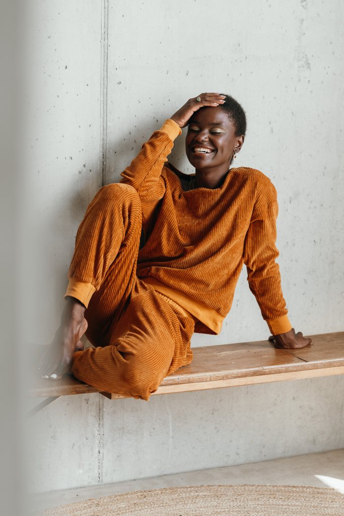 Model trägt ockerfarbigen Pyjama. Sie sitzt auf einer Sitzbank, die Beine auf der Bank angewinkelt und stützt den Kopf auf dem Ellbogen ab.