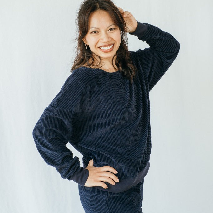 Model trägt Pyjama in Dunkelblauem Cord. Sie steht seitwärts, eine Hand im Haar und blickt direkt in die Kamera