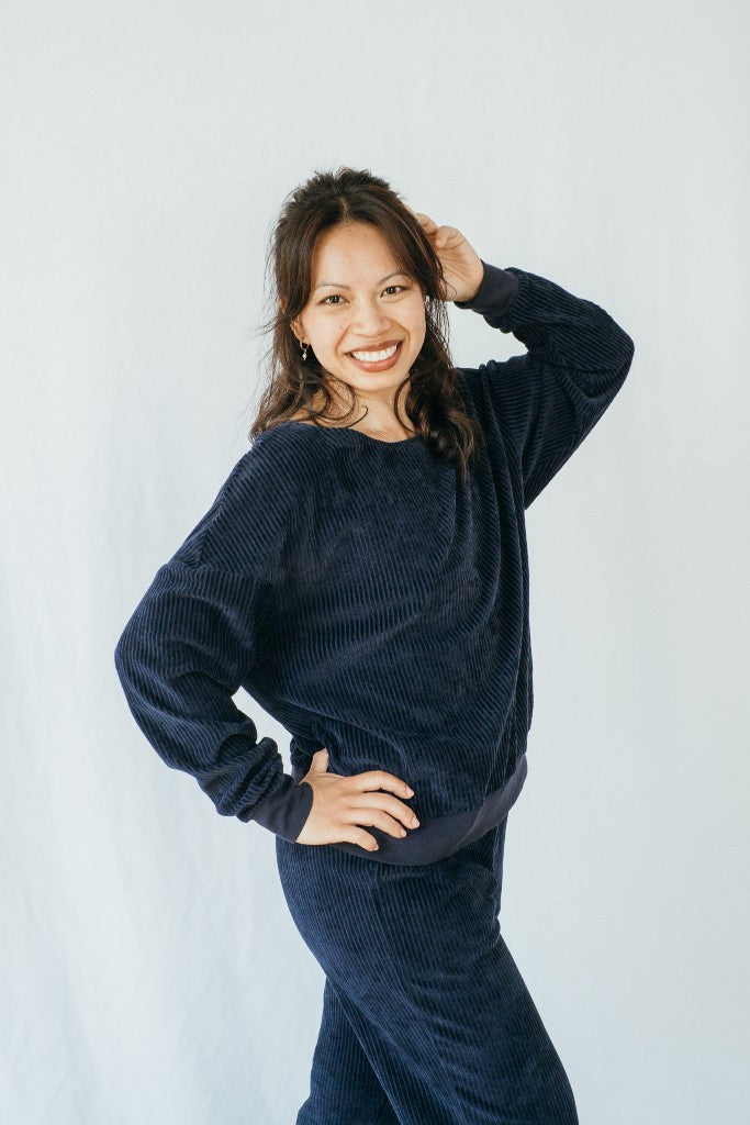 Model trägt Pyjama in Dunkelblauem Cord. Sie steht seitwärts, eine Hand im Haar und blickt direkt in die Kamera