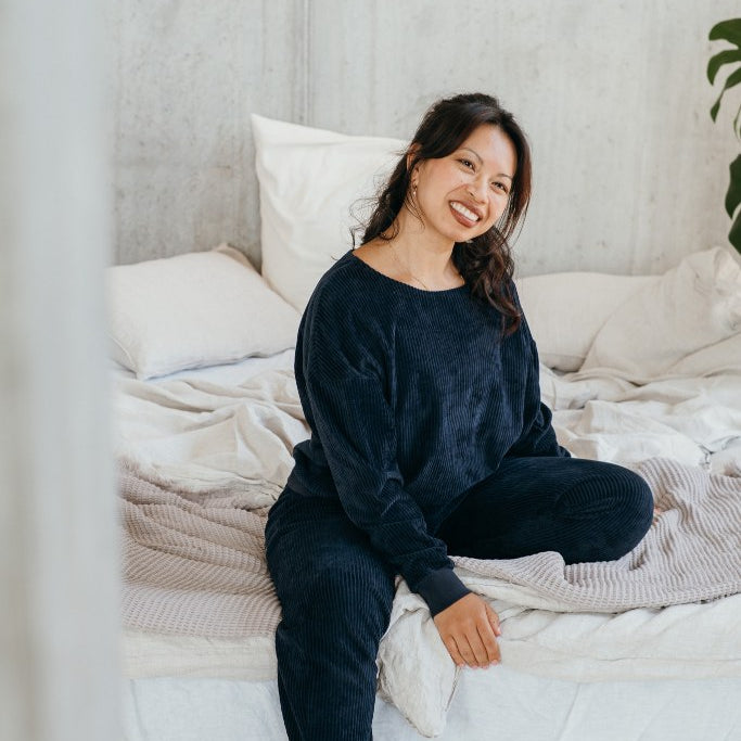 Model trägt Pyjama in Dunkelblauem Cord und sitzt auf dem Bett.
