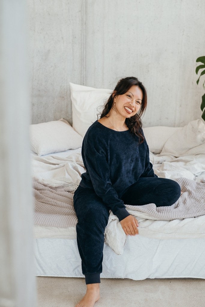 Model trägt Pyjama in Dunkelblauem Cord und sitzt auf dem Bett.