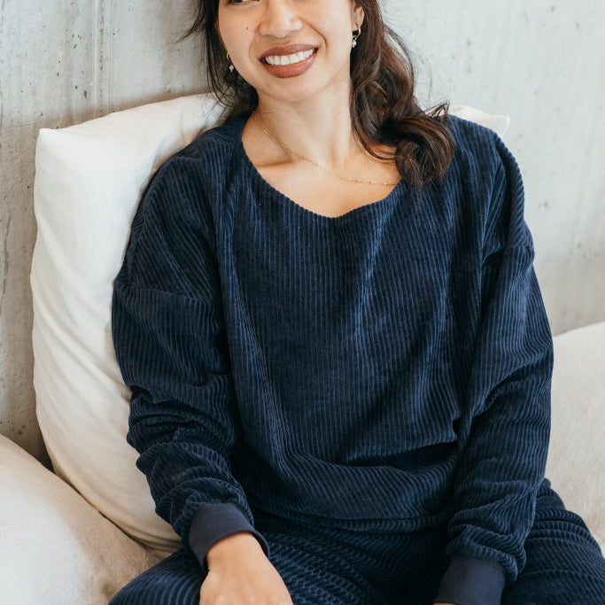 Model sitzt im dunkelblauen Pyjama auf dem Bett. Sie lehnt ihren Rücken an der Wand an und lächelt.