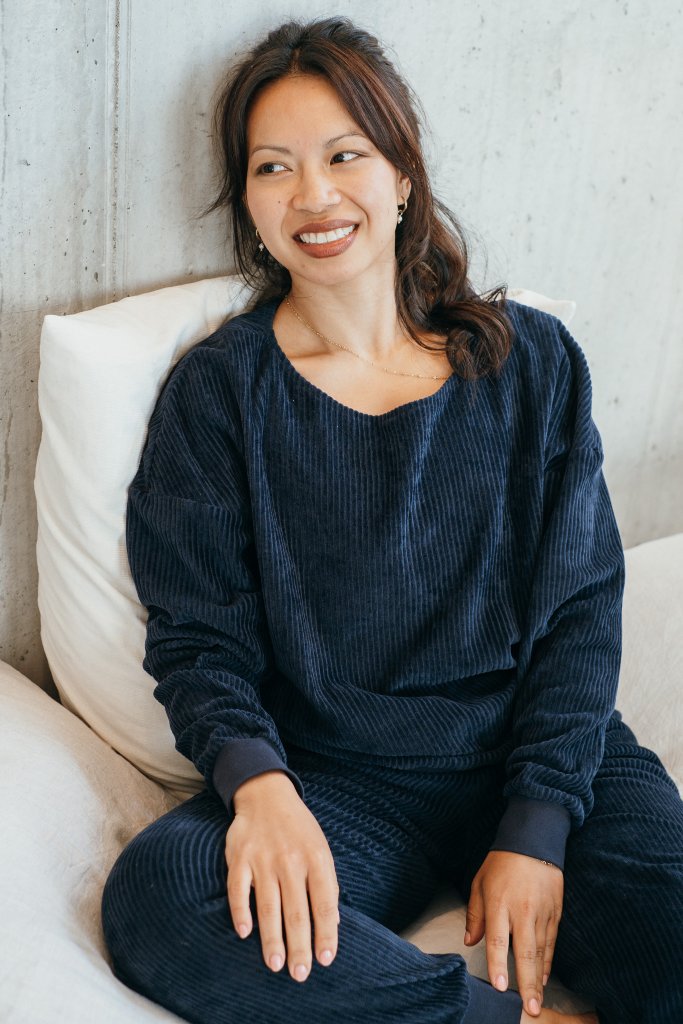 Model sitzt im dunkelblauen Pyjama auf dem Bett. Sie lehnt ihren Rücken an der Wand an und lächelt.