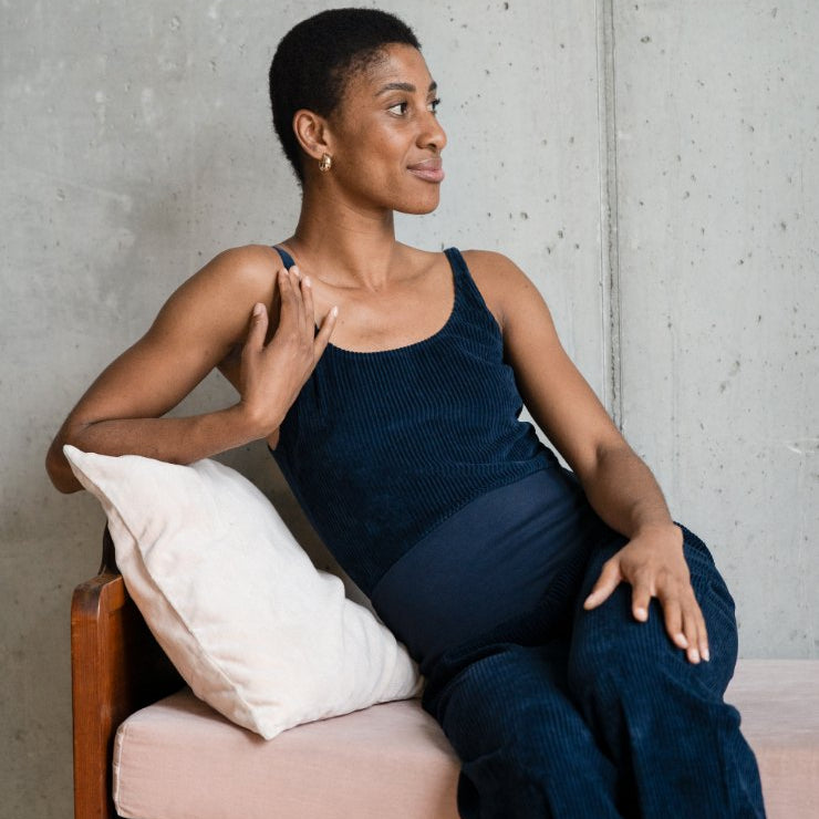 Model sitzt mit dem dunkelblauen Body und der Fiona Hose auf dem Tagesbett und blickt in Richtung des Fensters