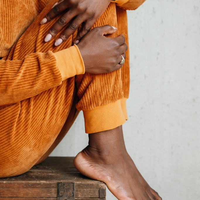 Detailaufnahme der Kelly Hose in Vibrant Ochre. Das Model sitzt auf einer Truhe und umschlingt ihre Beine mit beiden Armen.