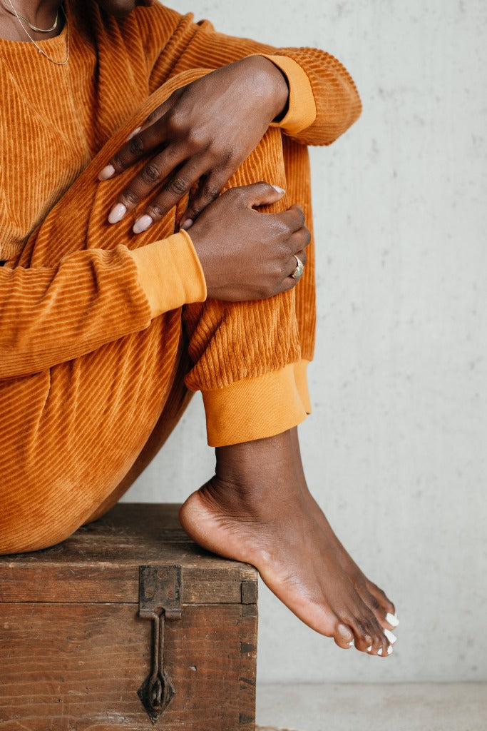 Detailaufnahme der Kelly Hose in Vibrant Ochre. Das Model sitzt auf einer Truhe und umschlingt ihre Beine mit beiden Armen.