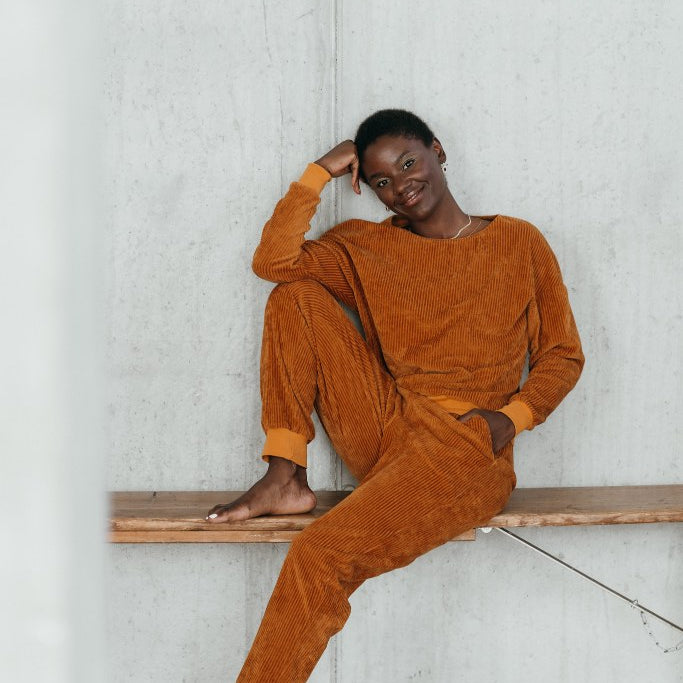 Model sitzt im ockerfarbigen Pyjama auf einer Sitzbank und lächelt direkt in die Kamera