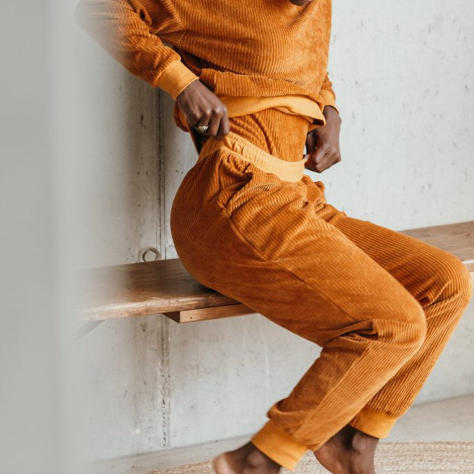 Model trägt den Pyjama aus ockerfarbigem Cord. Sie sitzt auf einer Sitzbank und zupft am Bund