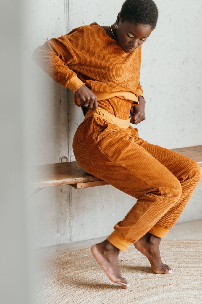 Model trägt den Pyjama aus ockerfarbigem Cord. Sie sitzt auf einer Sitzbank und zupft am Bund