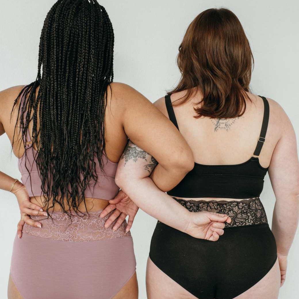 Deux femmes se tiennent en sous-vêtements, dos à la caméra, les bras accrochés.