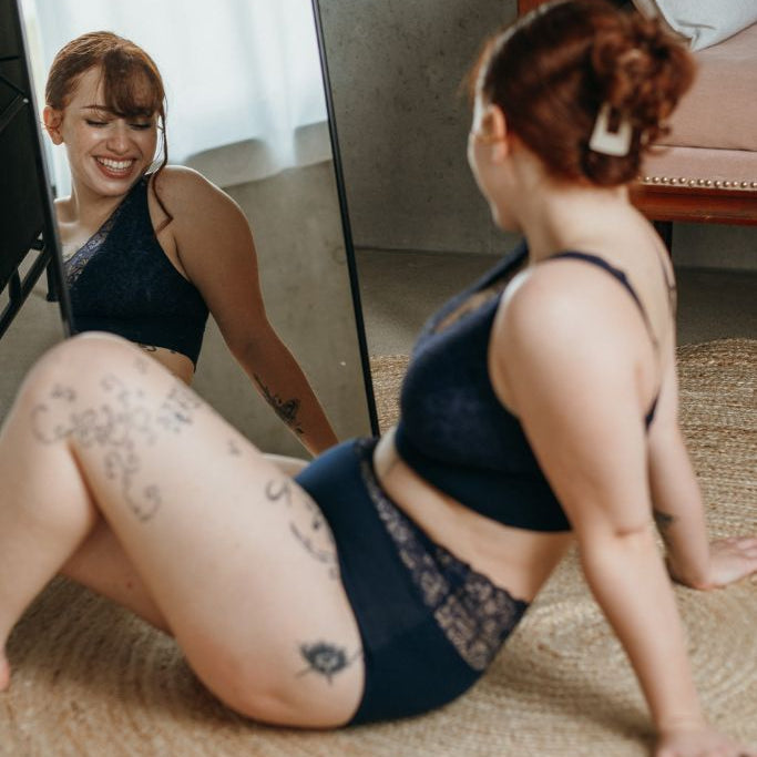 das lachende Model sitzt am Boden und betrachtet ihre Unterwäsche im Spiegel