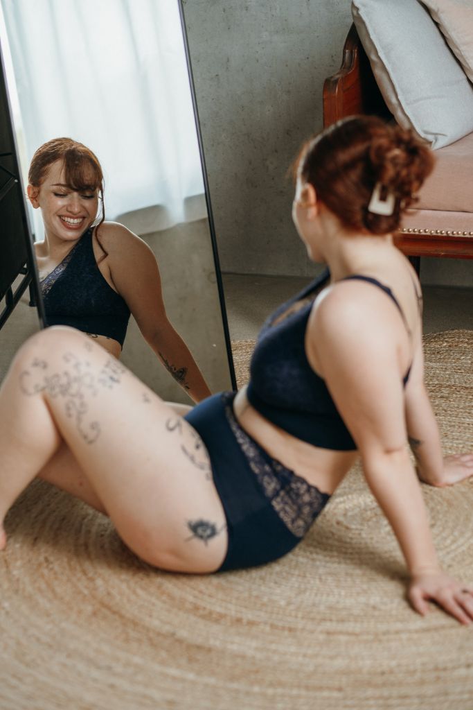 das lachende Model sitzt am Boden und betrachtet ihre Unterwäsche im Spiegel