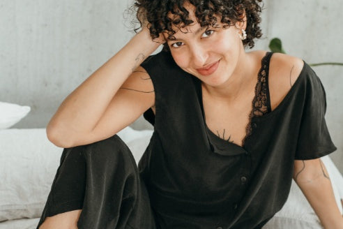 Le mannequin est assis sur le lit et présente le chemisier Noe et le pantalon Coco