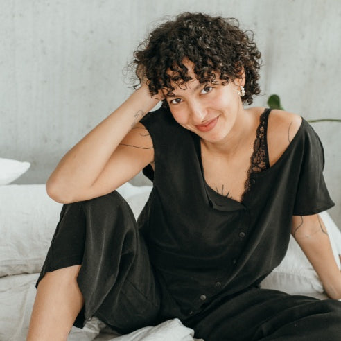 Le mannequin est assis sur le lit et présente le chemisier Noe et le pantalon Coco