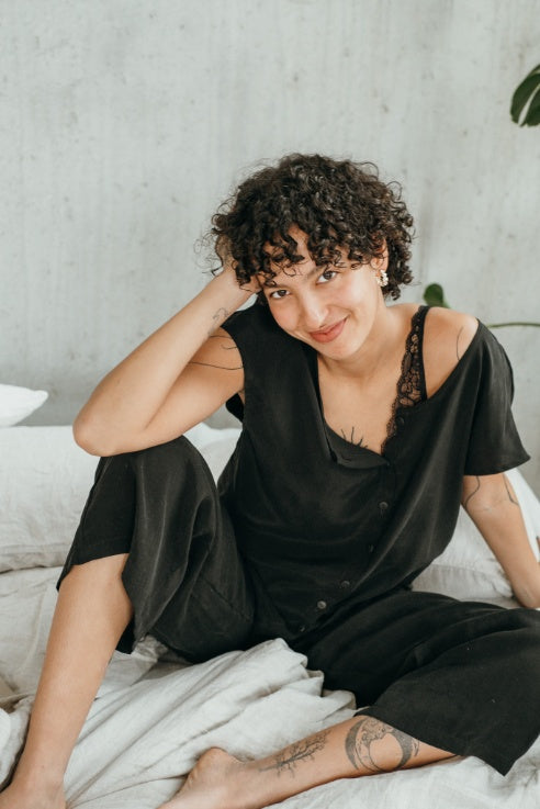 Le mannequin est assis sur le lit et présente le chemisier Noe et le pantalon Coco