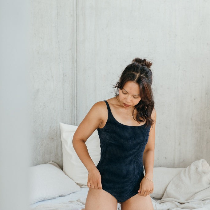 Model kniet auf dem Bett und trägt das dunkelblauen Ronja Body im Cord. Sie blickt an sich hinunter und zupft die Beinausschnitte zurecht.