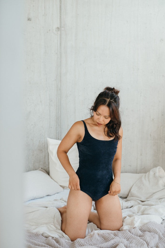 Model kniet auf dem Bett und trägt das dunkelblauen Ronja Body im Cord. Sie blickt an sich hinunter und zupft die Beinausschnitte zurecht.
