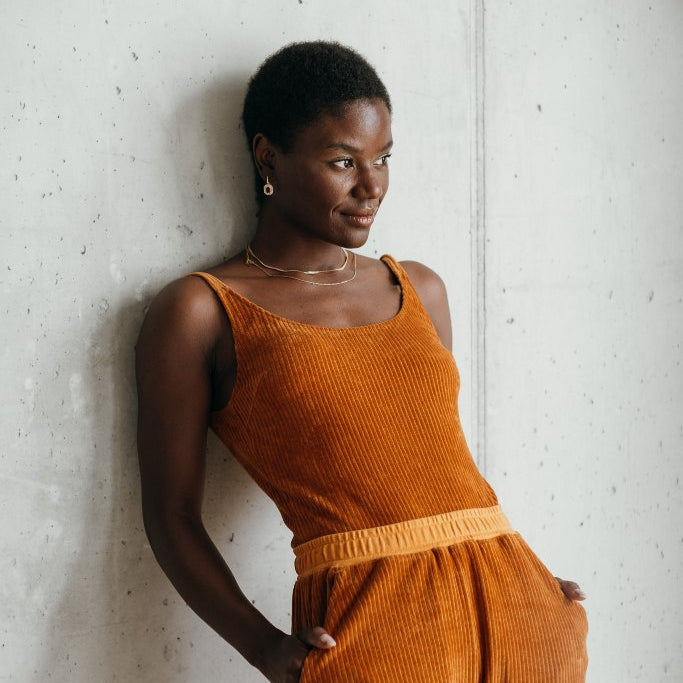 Model trägt ockerfarbiges Ronja Body und die Kelly Hose, sie lehnt entspannt an der Wand.