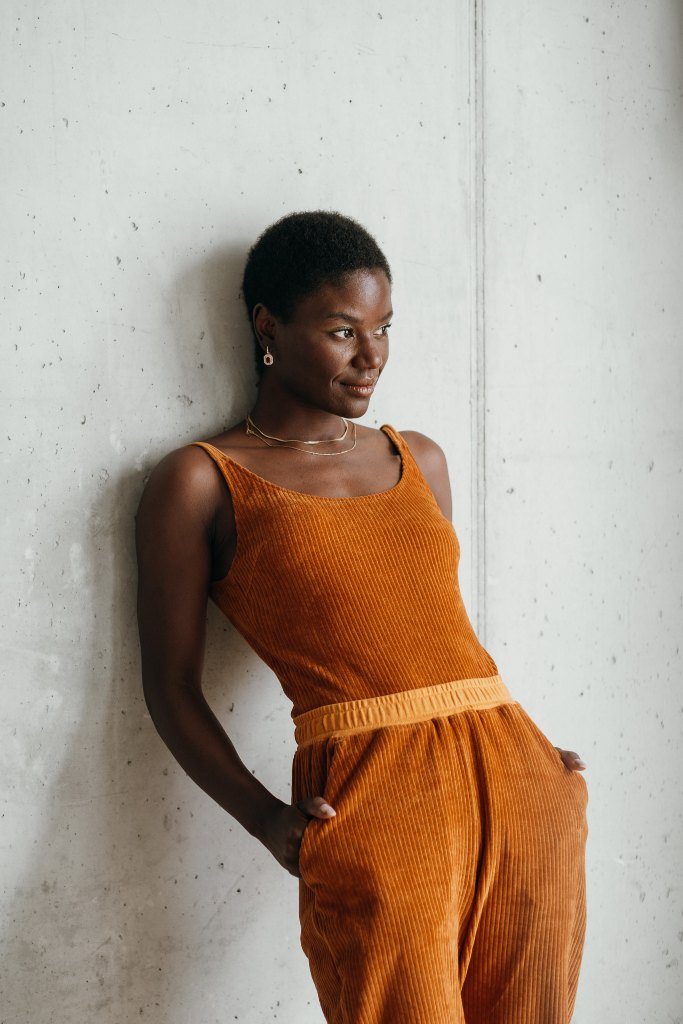 Model trägt ockerfarbiges Ronja Body und die Kelly Hose, sie lehnt entspannt an der Wand.