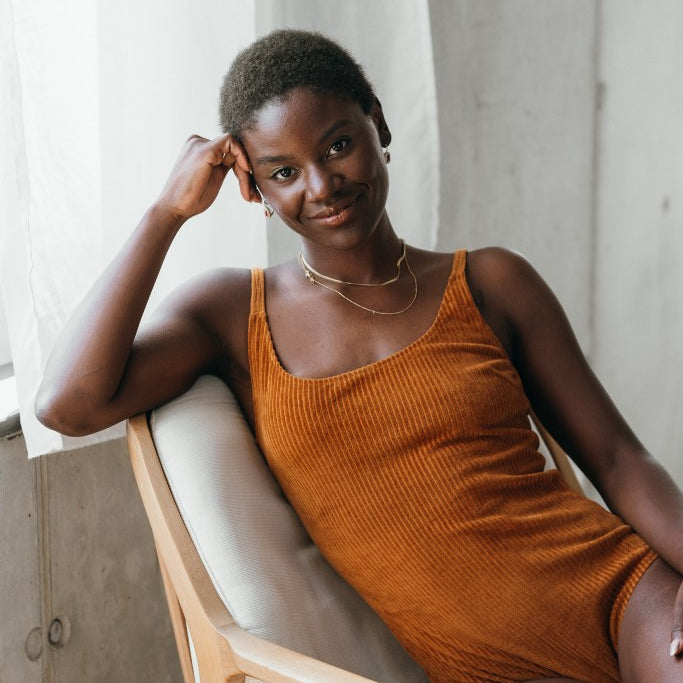 Model sitzt im ockerfarbigen Ronja Body auf einem weissen Sessel. Sie sieht direkt in die Kamera, den Kopf stützt sie auf einer Hand ab.