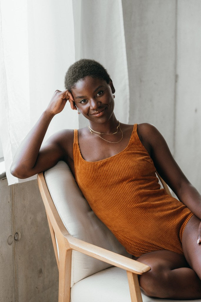 Model sitzt im ockerfarbigen Ronja Body auf einem weissen Sessel. Sie sieht direkt in die Kamera, den Kopf stützt sie auf einer Hand ab.