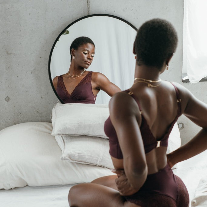 Model kniet auf dem Bett und blickt in den Spiegel. Sie trägt das Sophie Set in der Farbe Aubergine.