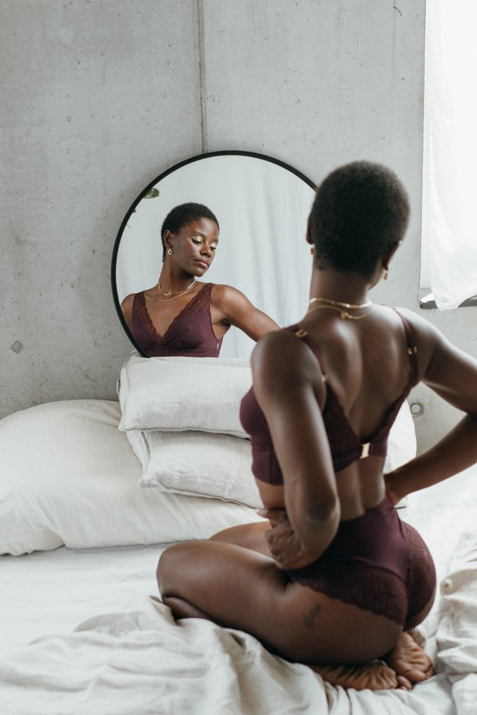 Model kniet auf dem Bett und blickt in den Spiegel. Sie trägt das Sophie Set in der Farbe Aubergine.