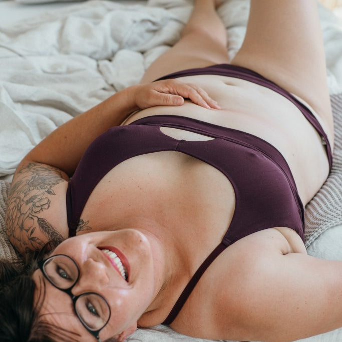 Model trägt das Thea Set in der Farbe Aubergine und liegt rücklings im Bett, ein Bein angewinkelt. Den Kopf streckt sie in die Kamera und lacht.