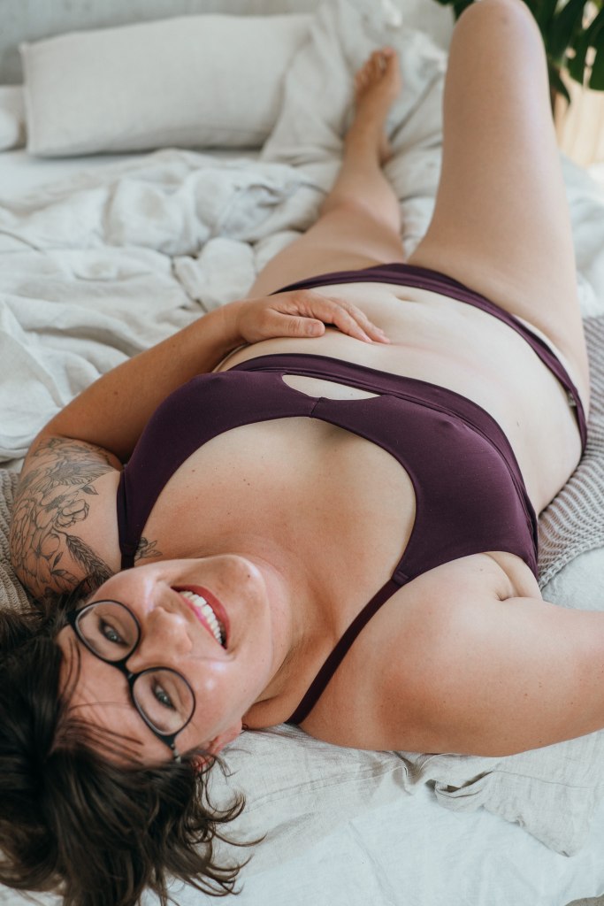 Model trägt das Thea Set in der Farbe Aubergine und liegt rücklings im Bett, ein Bein angewinkelt. Den Kopf streckt sie in die Kamera und lacht.