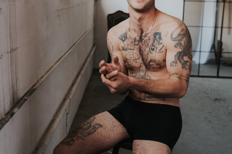 Menn est assis sur une chaise haute et rit à la caméra. Il porte un caleçon noir en TENCEL™