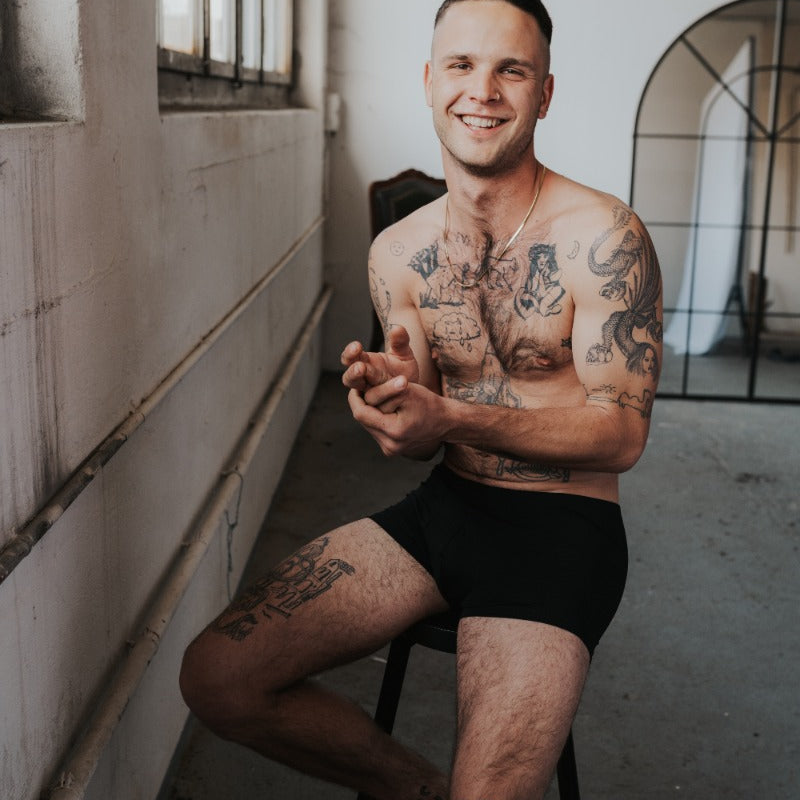 Menn est assis sur une chaise haute et rit à la caméra. Il porte un caleçon noir en TENCEL™