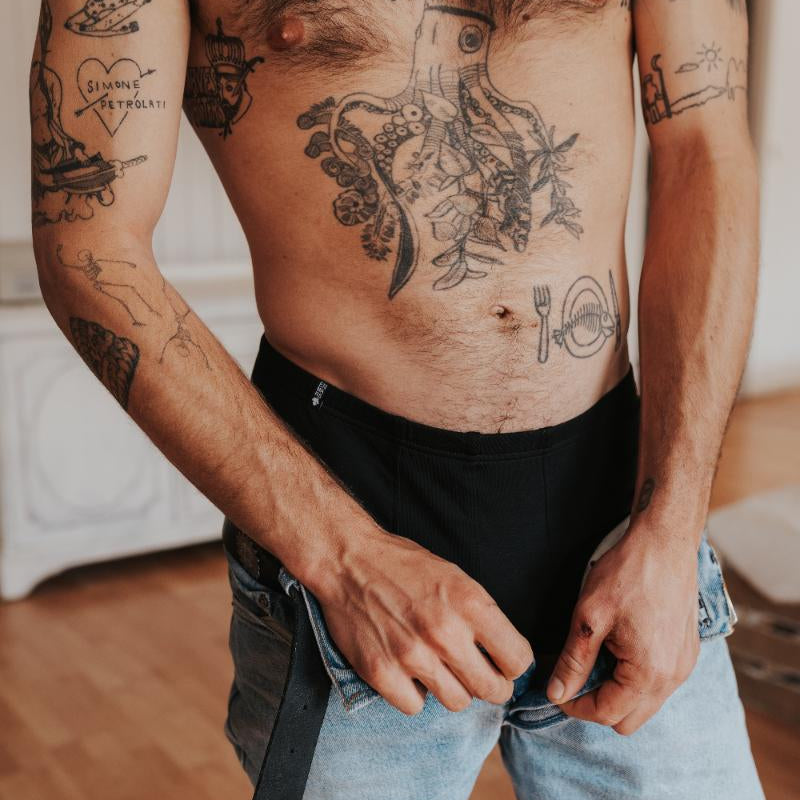 Un homme avec des tatouages enfile son pantalon, en dessous on voit un boxer noir.