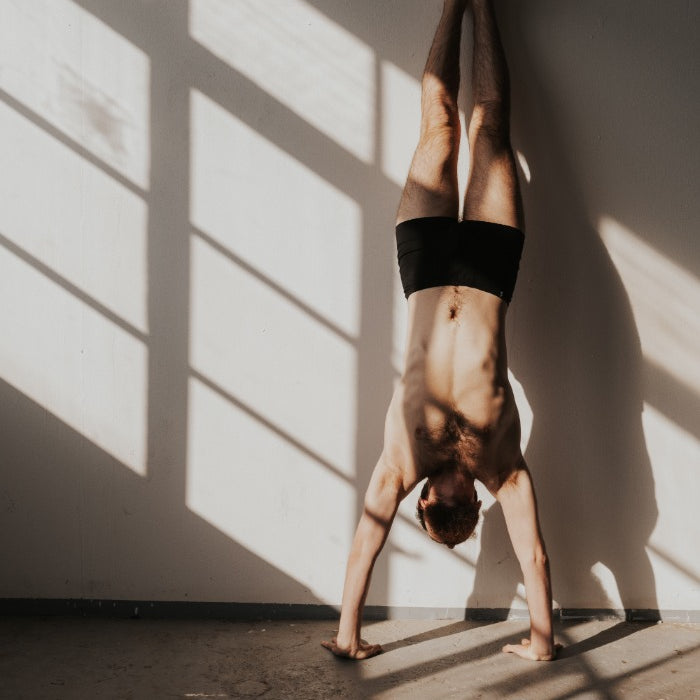 Une personne fait le poirier en boxer contre un mur.