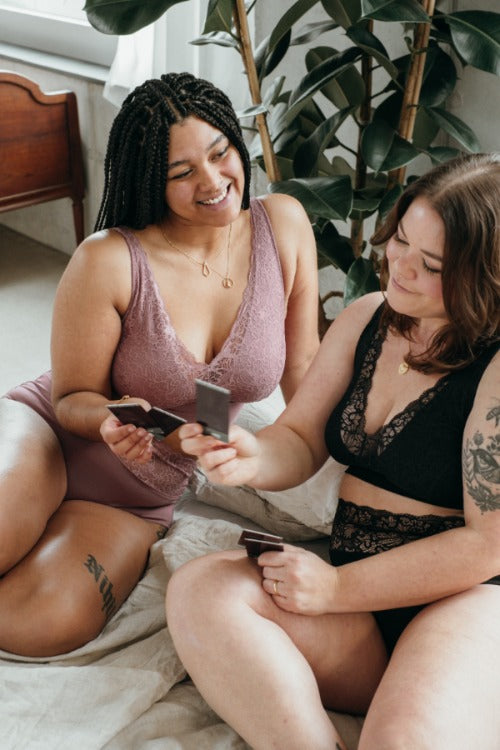 Two women in their underwear sit on a white bedspread and look at photos.