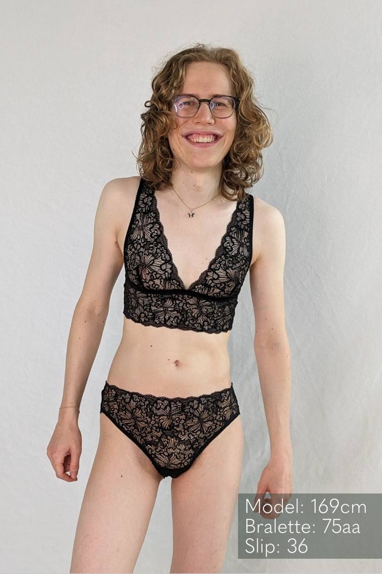 Woman stands in front of a white background in black Bralette and slip made of lace.