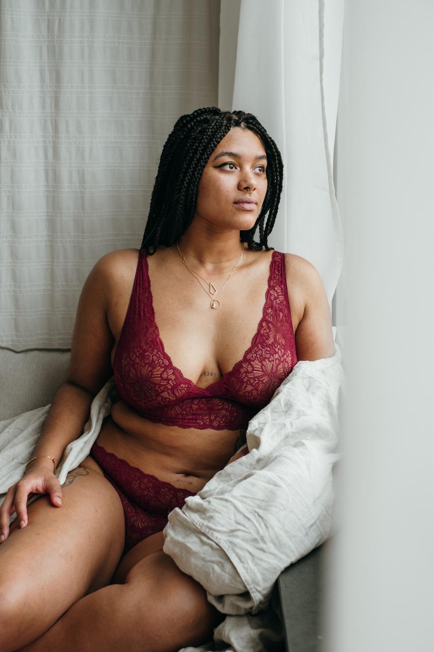 Model sits snuggled up in blanket in front of window and wears red lingerie.