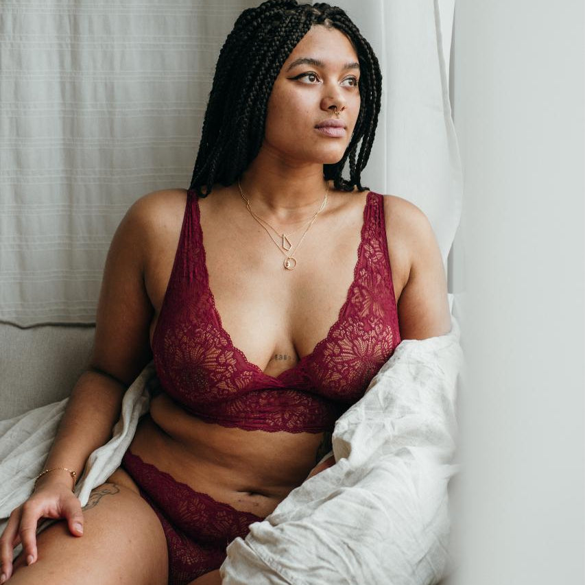 Model sits snuggled up in blanket in front of window and wears red lingerie.