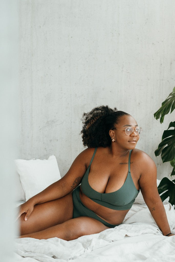 Model in Bralette and slip (dark green) sitting sideways on bed.