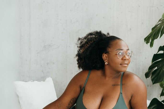 Model in Bralette und Slip (dunkelgrün) sitzt seitlich auf Bett.