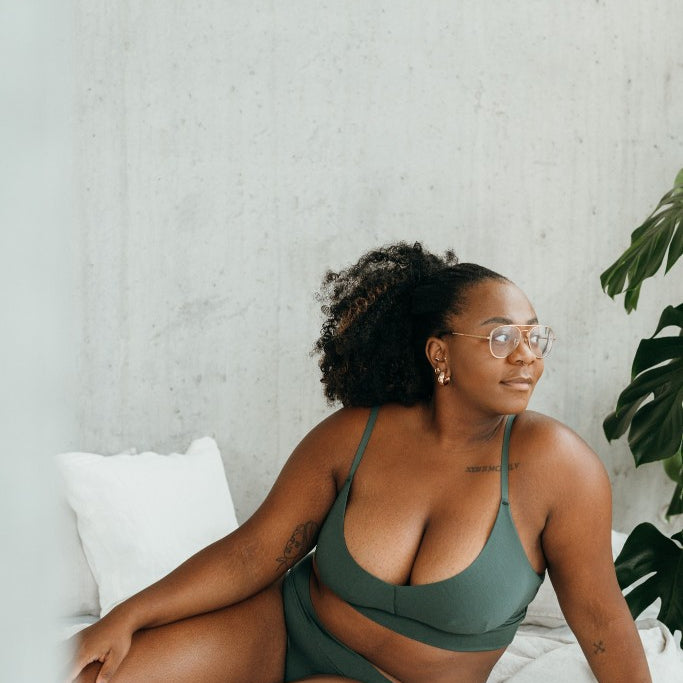 Model in Bralette und Slip (dunkelgrün) sitzt seitlich auf Bett.