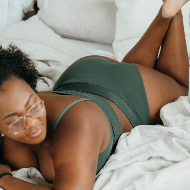 Model in grünem Dessous liegt auf Bett, der Fokus liegt auf dem feinen Slip.