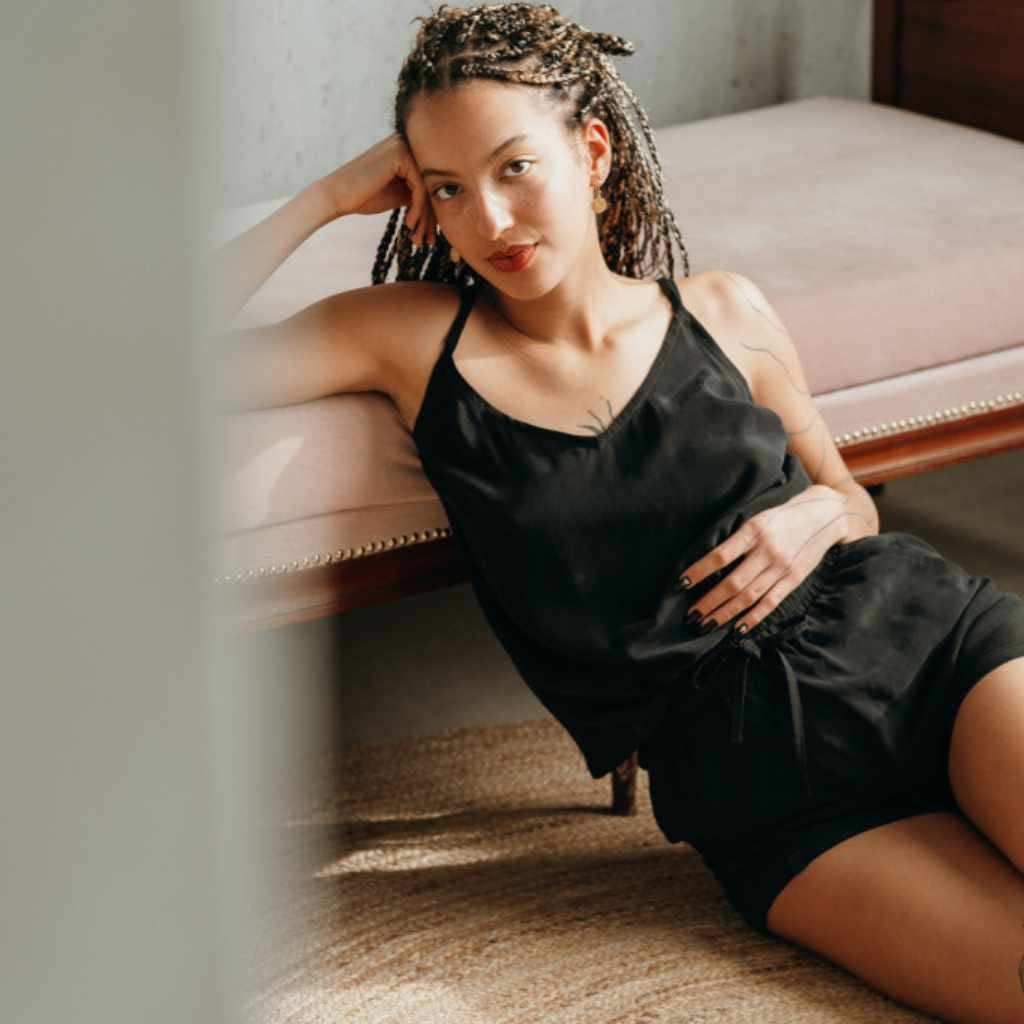 Model lehnt an rosa Sofa und trägt schwarzes Träger-Top und Shorts Kat aus Cupro.